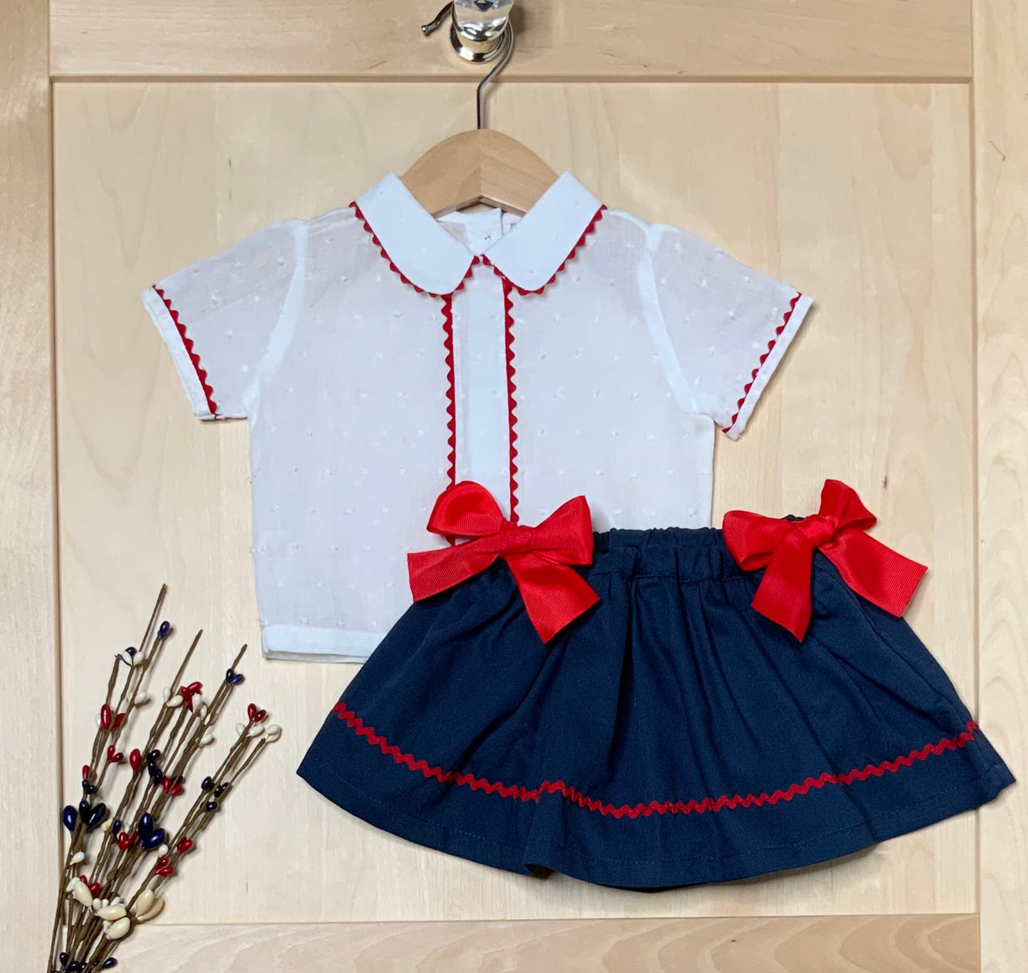 A toddler girl's two-piece outfit hanging on a wooden hanger against a light wood paneled background. The set includes a white short-sleeve collared top with red zigzag trim and a navy blue pleated skirt with red zigzag trim and large red bows at the waist-Alfa Baby Boutique 