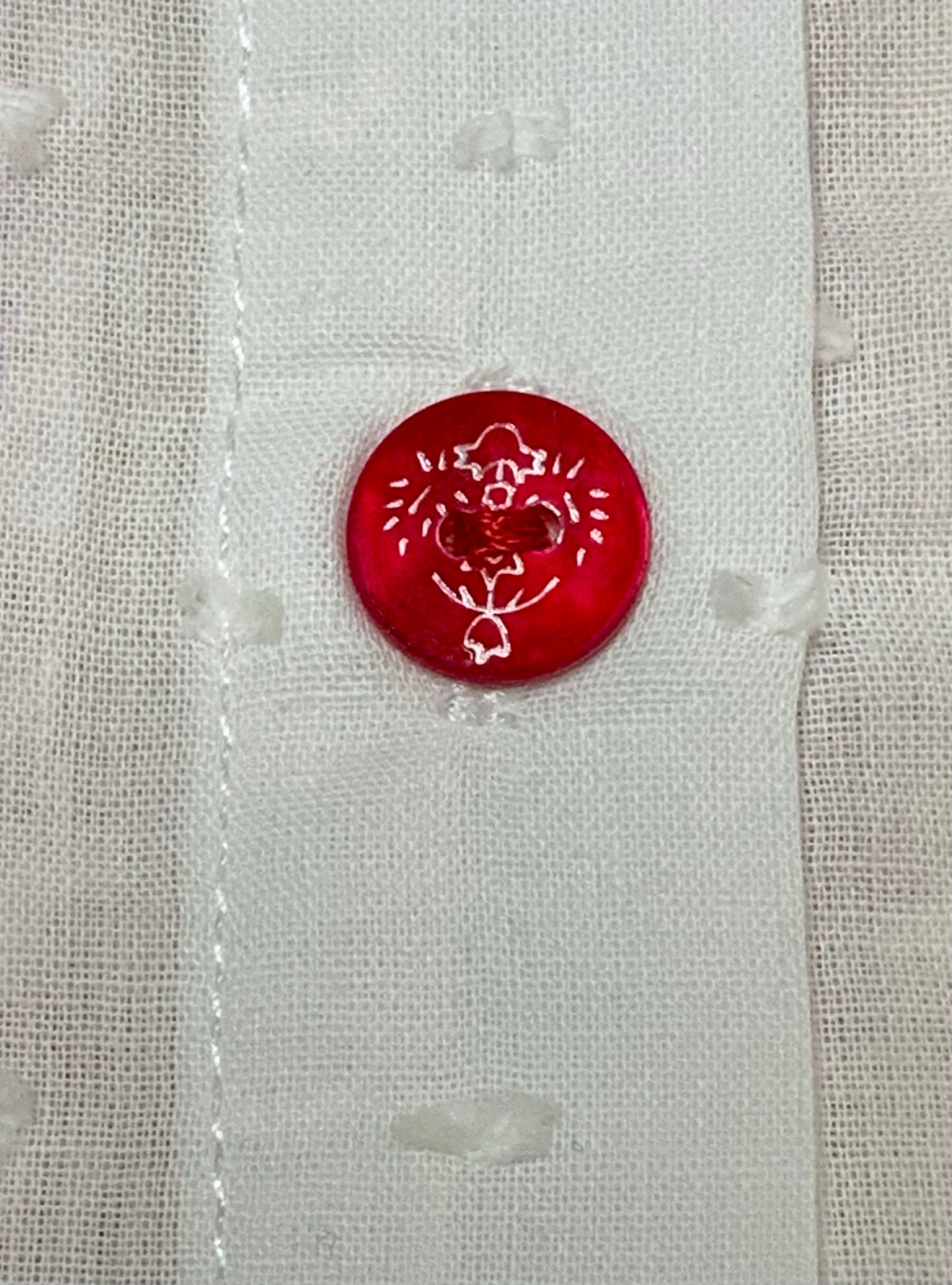 A close-up of a white textured plumeti fabric featuring a single round red button with white floral details-Alfa Baby Boutique 