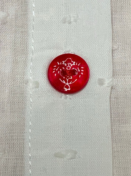 A close-up of a white textured plumeti fabric featuring a single round red button with white floral details-Alfa Baby Boutique 
