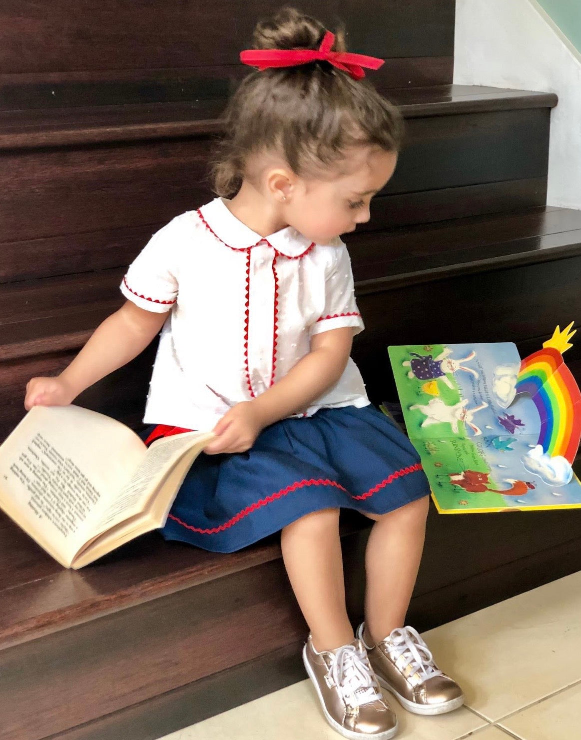 A toddler girl sits on dark wooden stairs, reading a book and holding a colorful children's picture book. She is wearing a white short-sleeve collared shirt with red trim, a navy blue skirt-Alfa Baby Boutique